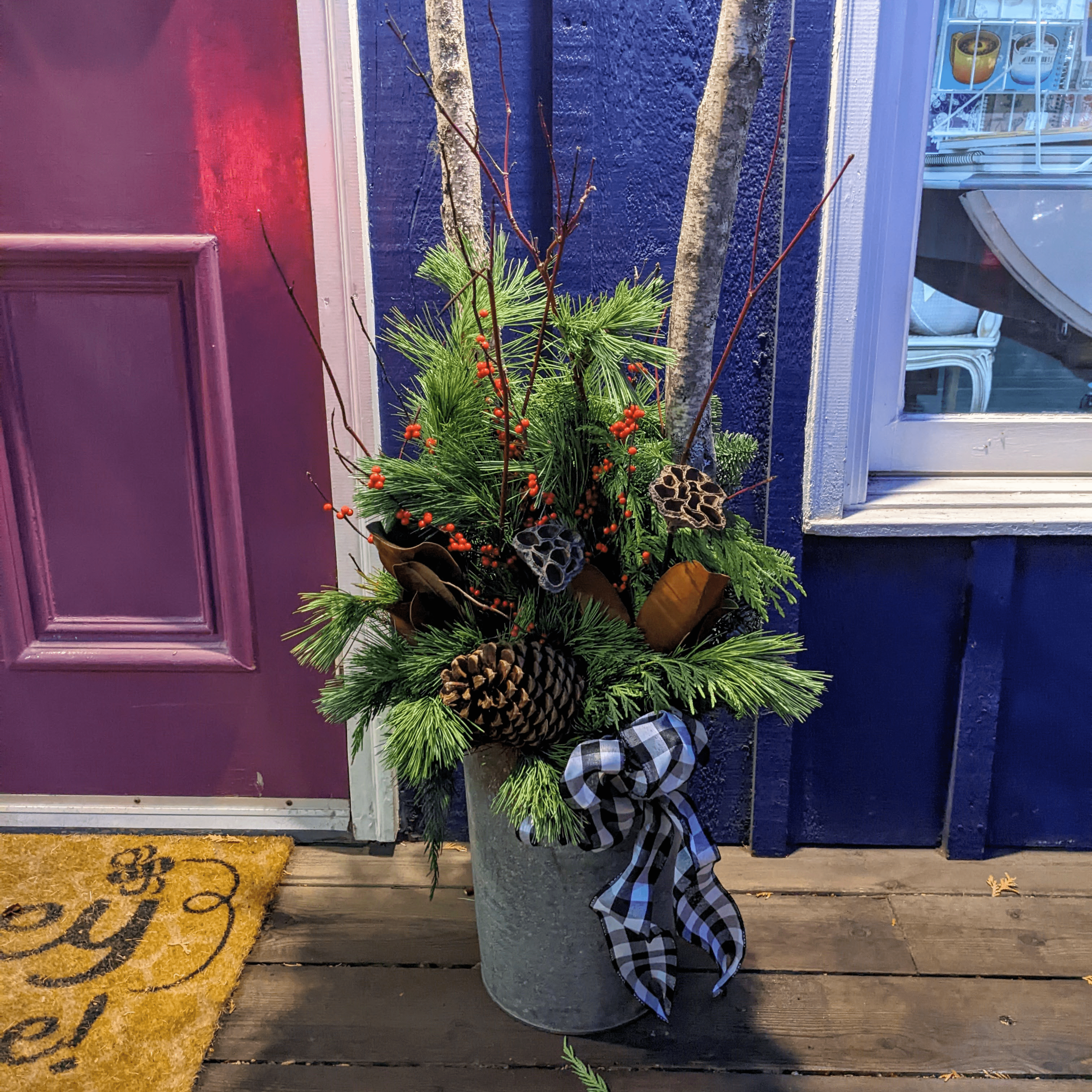 Winter Planter designed in a vintage metal sap bucket filled with a mix of greenery, fresh red berries, pine cones, magnolia leaves, red dog wood and birch sticks sitting in front of a purple door and blue wall book shop.