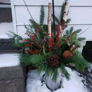 Winter Planter designed in a vintage metal sap bucket filled with a mix of greenery, fresh red berries, pine cones, magnolia leaves, red dog wood and birch sticks sitting in front of a white siding house.